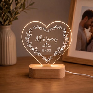 Op de afbeelding: Een hartvormige acryl lamp met een houten voet. Het acryl is voorzien van een gegraveerd bloemenkransontwerp, de namen Milli & Laurenz, een afbeelding van twee in elkaar grijpende ringen en de datum 08.08.2028, verlicht door een warme gloed.