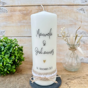 May include: White pillar candle with the names "Manuela & Johannes" and the date "04. NOVEMBER 2023" written on it. The candle is decorated with a small gold heart and a brown ribbon.