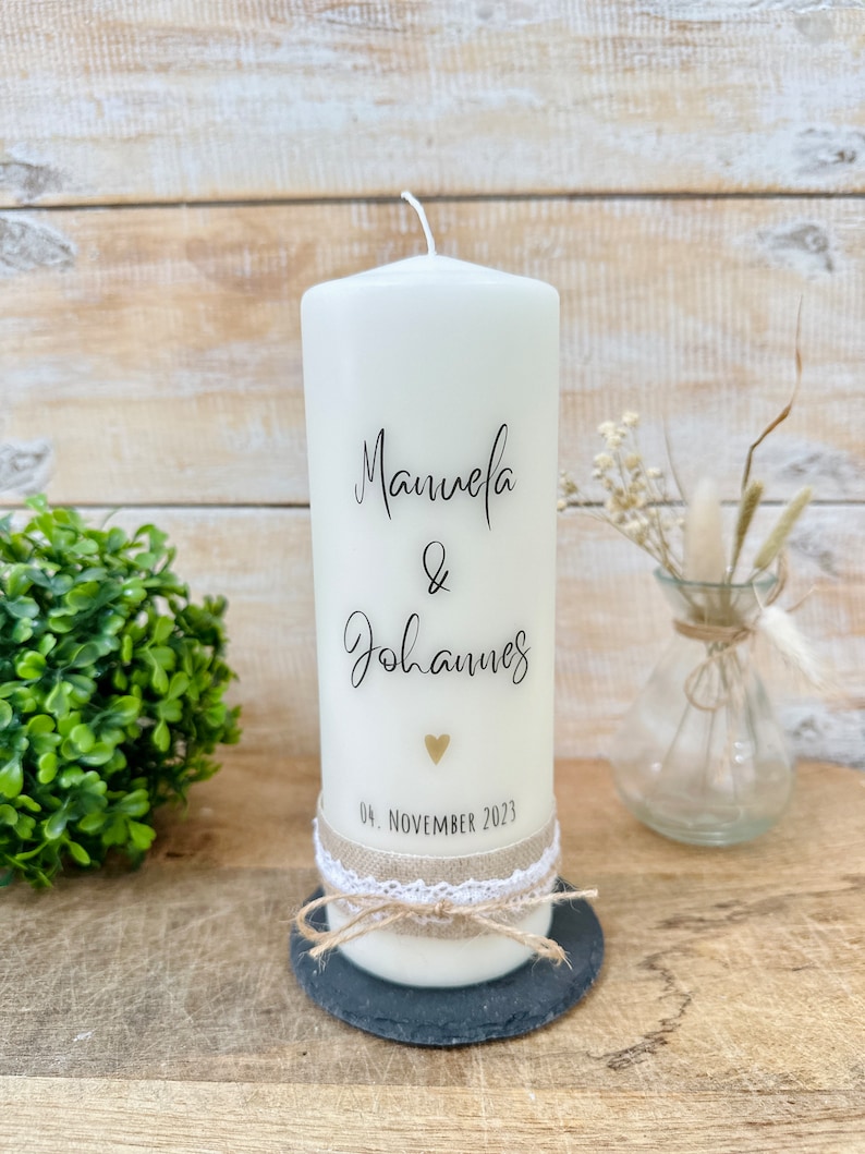 May include: White pillar candle with the names "Manuela & Johannes" and the date "04. NOVEMBER 2023" written on it. The candle is decorated with a small gold heart and a brown ribbon.