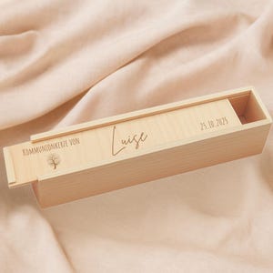 Puede incluir: Una caja de madera de color claro con tapa corredera. La tapa está grabada con el nombre "Luise" en cursiva, la fecha "25.10.2025" y las palabras "KOMMUNIONKERZE VON" con un gráfico de árbol. La caja está abierta, revelando un interior vacío.
