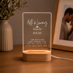 Puede incluir: Un panel de luz acrílico rectangular con base de madera, que presenta los nombres Milli & Laurenz, un gráfico de anillos de boda, la fecha 08.08.2028 y una cita en alemán sobre la fe, la esperanza y el amor, iluminado con un brillo blanco cálido.