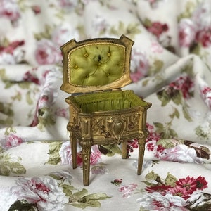 May include: A small, ornate, gold-colored jewelry box with a green velvet interior. The box is on four legs and has a hinged lid.