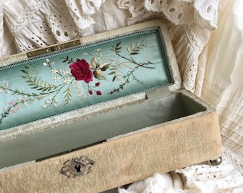 May include: An antique jewelry box with a light blue velvet interior and a floral embroidery design. The box is made of beige velvet and has a silver lock and hinges.