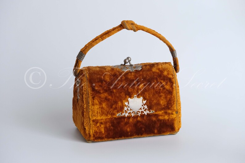 May include: A vintage velvet handbag with a knotted handle and a silver clasp. The bag is a rich brown color and has a silver shield design on the front.