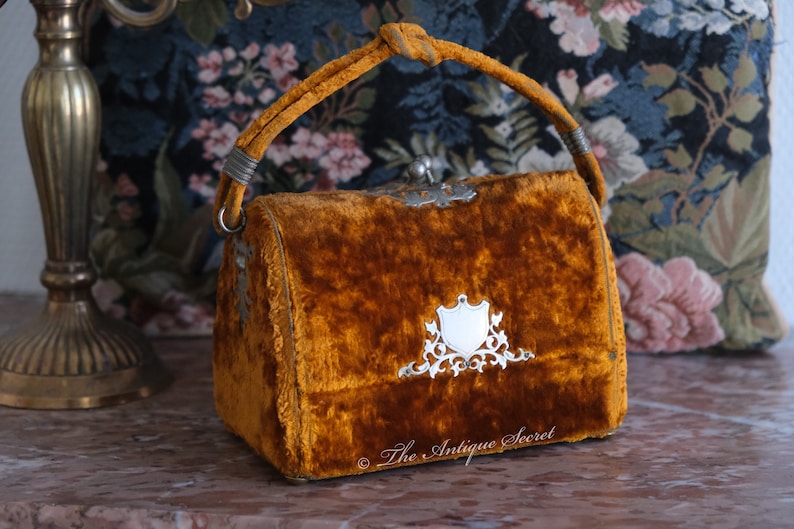 May include: A small, vintage, mustard yellow velvet handbag with a silver clasp and ornate silver design. The bag has a top handle and is in good condition.