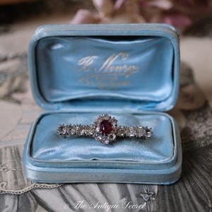 Puede incluir: Broche vintage con un rubí central ovalado rodeado de piedras claras, presentado en una caja de terciopelo azul claro. La caja tiene letras doradas. El broche se encuentra sobre un interior de terciopelo azul claro.