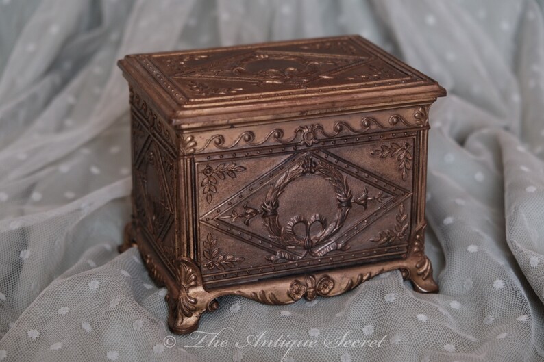Puede incluir: Una peque&ntilde;a caja de madera adornada con intrincados tallados y un acabado dorado. La caja tiene una tapa con bisagras y se asienta sobre cuatro patas decorativas.