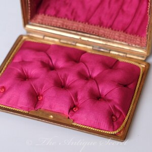 Puede incluir: Una peque&ntilde;a caja rectangular dorada con un interior de terciopelo rosa. La caja est&aacute; abierta y el forro de terciopelo es visible. La caja tiene un borde dorado alrededor del borde.