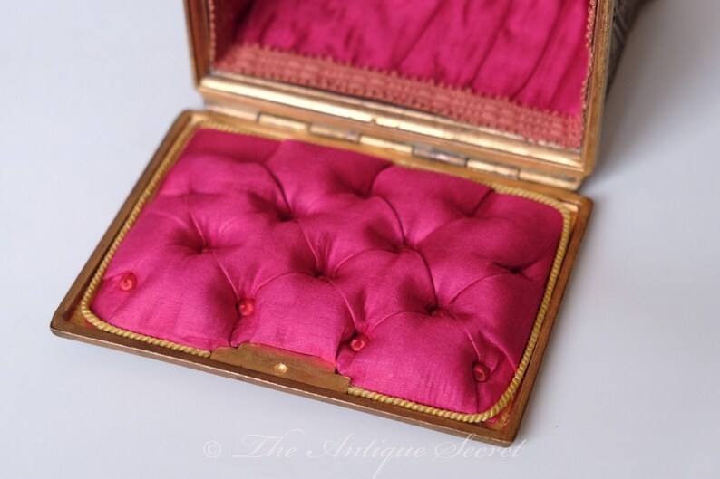 Puede incluir: Una peque&ntilde;a caja rectangular dorada con un interior de terciopelo rosa. La caja est&aacute; abierta y el forro de terciopelo es visible. La caja tiene un borde dorado alrededor del borde.