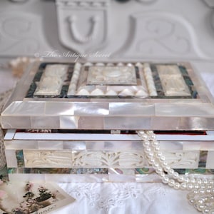 Puede incluir: Una caja de joyería de nácar blanco con diseños intrincados. La caja está hecha de pequeñas baldosas iridiscentes y tiene una tapa con bisagras. La caja está abierta y una hebra de perlas blancas se derrama.