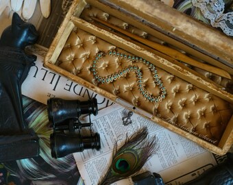 May include: An antique wooden box with a gold satin lining and a turquoise beaded necklace inside. The box is open and the necklace is visible. The box is sitting on a table with a newspaper and a peacock feather.