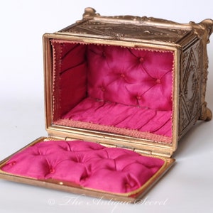Puede incluir: Una caja de joyer&iacute;a de metal dorado adornada, con una tapa con bisagras y un interior de terciopelo rosa. La caja tiene tallas intrincadas y un dise&ntilde;o en relieve.