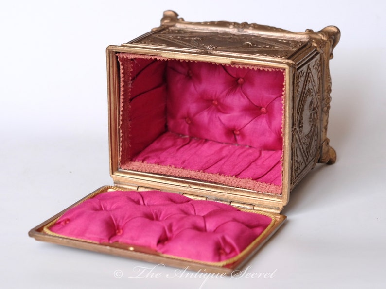 Puede incluir: Una caja de joyer&iacute;a de metal dorado adornada, con una tapa con bisagras y un interior de terciopelo rosa. La caja tiene tallas intrincadas y un dise&ntilde;o en relieve.