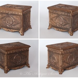 Puede incluir: Una peque&ntilde;a caja de madera antigua adornada con una tapa con bisagras. La caja est&aacute; hecha de madera oscura y tiene tallas intrincadas en la parte delantera y los lados. La caja est&aacute; decorada con un acabado dorado y tiene un dise&ntilde;o decorativo en la tapa.