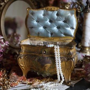 May include: An ornate gold-toned jewelry box with a blue velvet interior. The box is open and features a pearl necklace and a ring inside. The box is sitting on a table with a floral pattern.