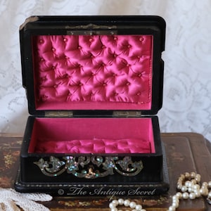 May include: An antique wooden jewellery box with a black finish and a pink satin lining. The box is decorated with a floral design and has a small metal latch. The box is open and the inside is visible. The box is sitting on a wooden table.