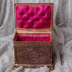 Puede incluir: Una caja de joyer&iacute;a antigua de color dorado con intrincados dise&ntilde;os florales y un panel central en forma de diamante. La caja est&aacute; abierta, revelando un interior de terciopelo rojo con acolchado de botones.