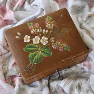 May include: A wooden box with a floral design painted on the lid. The flowers are white with green leaves and pink berries. The box has a keyhole and a small key attached to it. The text "The Antique Secret" is printed on the bottom right corner of the box.