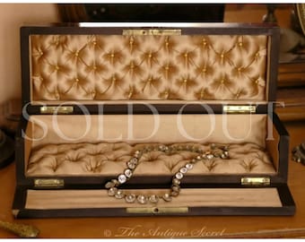 May include: A rectangular dark wood jewellery box with a hinged lid, featuring a tufted champagne-coloured satin interior lining with gold-toned button accents. A delicate necklace with circular clear stones rests inside the open box.