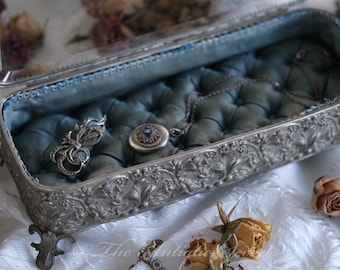 May include: An ornate silver jewellery box with a blue velvet lining. The box has a hinged lid with a mirror inside and sits on four decorative feet. The box is open and contains a silver necklace, a silver brooch, and a silver locket.