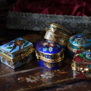 May include: A collection of small, ornate antique boxes. The boxes feature intricate designs with gold trim and vibrant colors, including blue, red, and gold. Each box has a unique floral or bird motif.