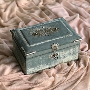May include: A vintage green velvet jewelry box with a silver metal latch and ornate design. The box has a keyhole and a tarnished silver metal design on the lid.