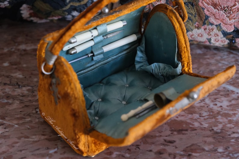 May include: An antique velvet vanity case with a blue satin lining. The case is open and reveals a set of silver-plated tools, including a small pair of scissors, a tweezers, and a nail file.