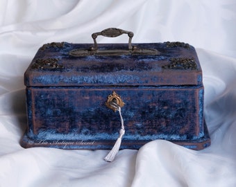 May include: A vintage blue velvet jewelry box with ornate metal hardware. The box features a handle, corner accents, and a keyhole with a tassel. The box is rectangular and has a distressed, aged appearance, suggesting it is an antique.