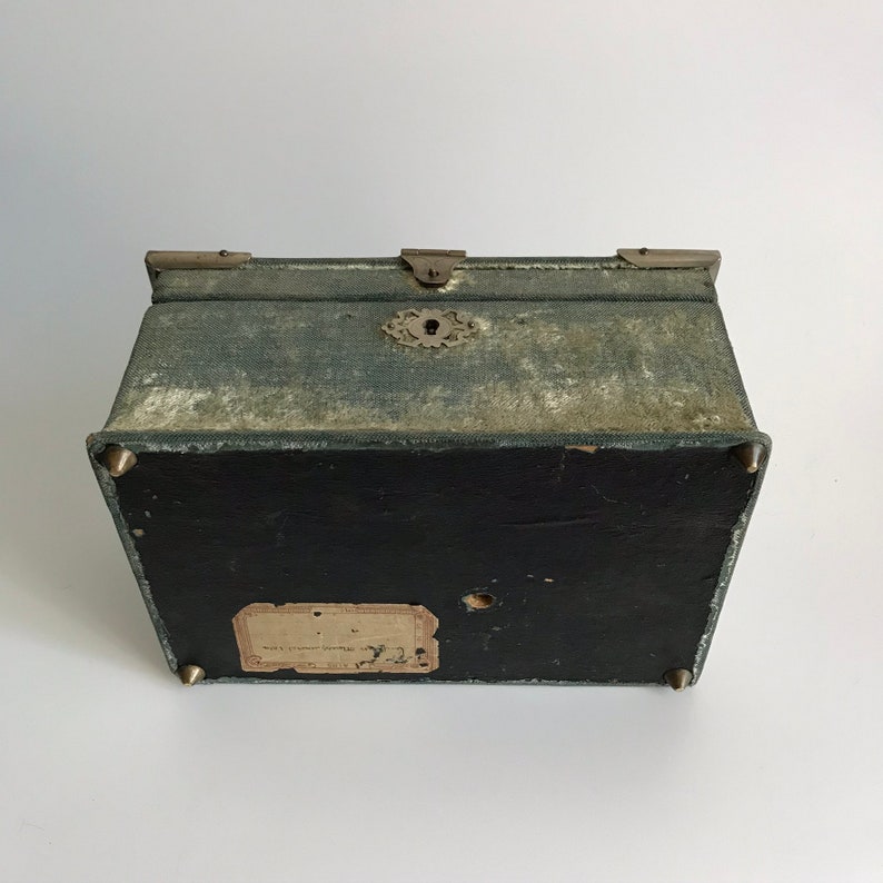 May include: A vintage green fabric covered box with brass hardware and a keyhole. The box has a label on the bottom that reads "Made in England by J.W. Benson Ltd."