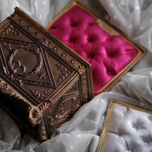 Puede incluir: Una caja de joyer&iacute;a vintage de color dorado adornada con un dise&ntilde;o floral detallado. La caja est&aacute; abierta y revela un interior de terciopelo rosa. La caja est&aacute; sentada sobre una tela de encaje blanco.