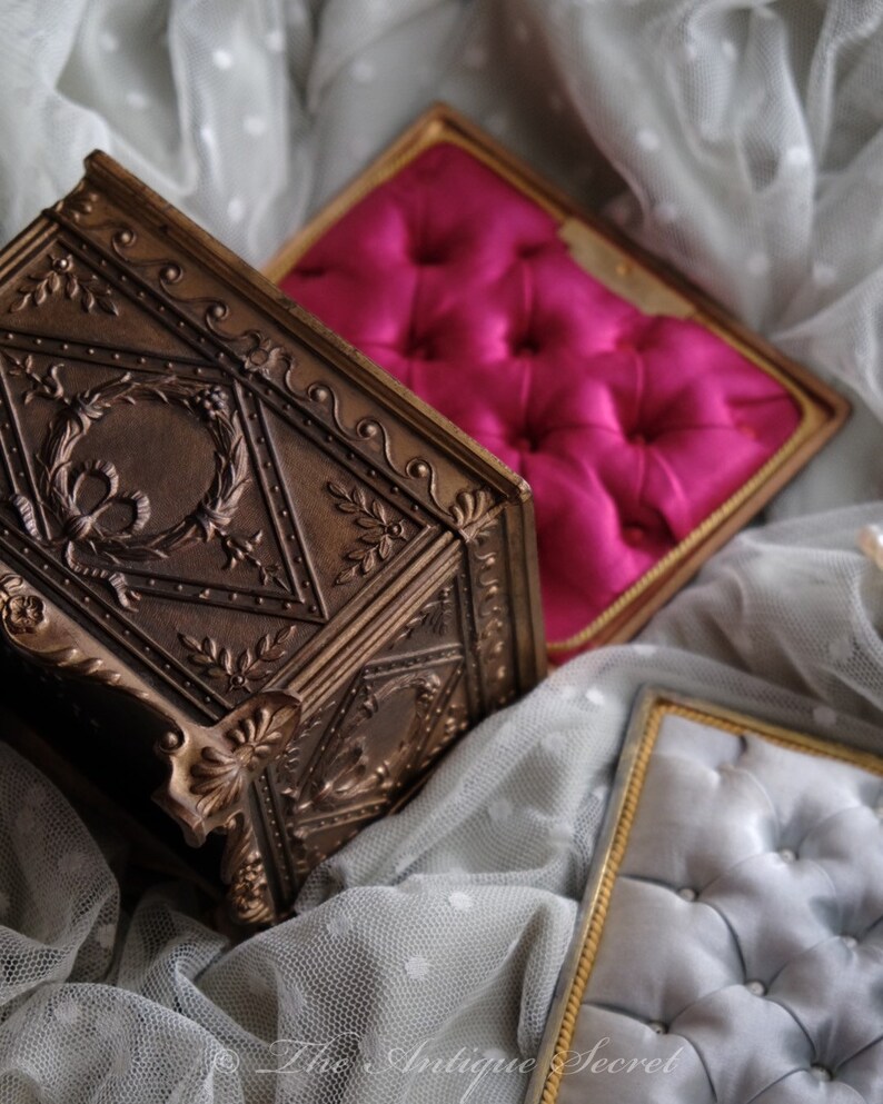 Puede incluir: Una caja de joyer&iacute;a vintage de color dorado adornada con un dise&ntilde;o floral detallado. La caja est&aacute; abierta y revela un interior de terciopelo rosa. La caja est&aacute; sentada sobre una tela de encaje blanco.