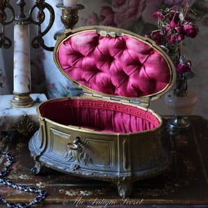 May include: An antique, oval-shaped jewelry box with a gold-toned metal exterior and a plush, pink interior. The box is open, revealing the quilted fabric lining. A decorative necklace rests nearby. The box is on a wooden surface.