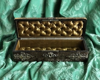 May include: An antique wooden box with a gold velvet interior and silver metal accents. The box is open and the interior is visible. The box is sitting on a green fabric surface.