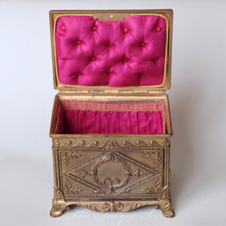 Puede incluir: Una caja de joyer&iacute;a antigua de tono dorado con un interior de terciopelo rosa. La caja tiene un dise&ntilde;o ornamentado con un motivo floral y una corona en relieve.