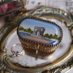 Op de afbeelding: Een klein, ovaalvormig, goudkleurig sieradendoosje met een scharnierend deksel. Het deksel is versierd met een geschilderd tafereel van de Arc de Triomphe in Parijs, Frankrijk. Het doosje staat op een gouden dienblad.