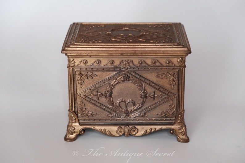Puede incluir: Una caja de madera de color dorado con tallas intrincadas y un dise&ntilde;o decorativo. La caja tiene una tapa con bisagras y se asienta sobre cuatro patas adornadas.