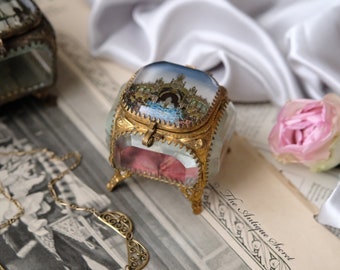 May include: Two antique glass trinket boxes with gold trim and painted scenes on the lids. The boxes are sitting on a newspaper with a gold chain necklace and a pink rose.