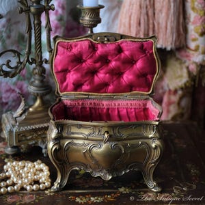 May include: An ornate gold-toned jewelry box with a red velvet interior. The box is open and has a decorative design. The box is sitting on a table with a pearl necklace.