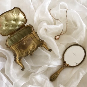 May include: A vintage gold-tone jewelry box with intricate floral details and a hinged lid. The box is open and has a white satin lining. A gold-tone hand mirror with a decorative handle is also pictured.