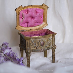 May include: An antique gold-tone jewelry box with a pink velvet interior and intricate carvings. The box is open and sits on a white fabric surface.
