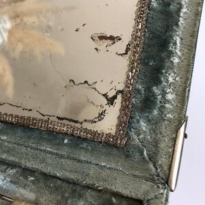 May include: Close-up of a vintage vanity mirror with a silver trim and a green velvet lining. The mirror is slightly damaged and has a faded appearance.