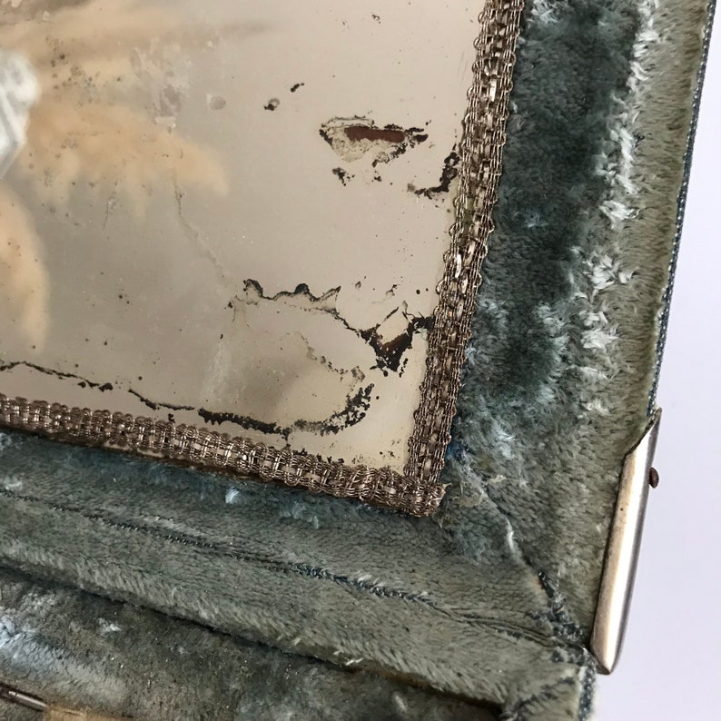May include: Close-up of a vintage vanity mirror with a silver trim and a green velvet lining. The mirror is slightly damaged and has a faded appearance.