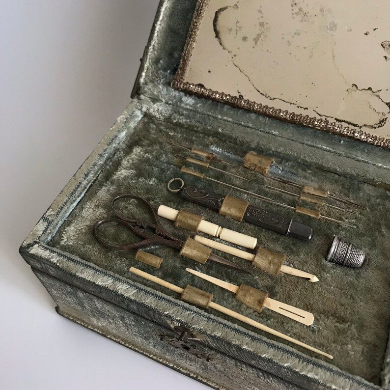 May include: An antique sewing kit with a green velvet lining. The kit includes a pair of scissors, a thimble, needles, and a small mirror.