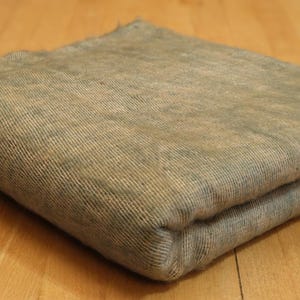 Puede incluir: Manta rectangular plegada en tonos grises y beige. La manta tiene una textura tejida y parece suave. Los bordes están ligeramente deshilachados. La manta está doblada cuidadosamente, mostrando las variaciones de textura y color.