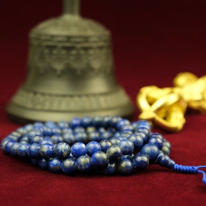 May include: A blue lapis lazuli mala prayer bead necklace with a tassel. The necklace is lying on a red surface with a golden bell and a golden vajra in the background.