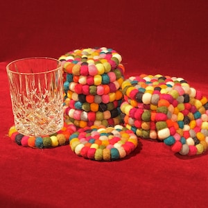 May include: A set of colourful felt coasters made of small, round balls. The coasters are stacked and arranged in a circular pattern. The coasters are a mix of bright colours, including red, orange, yellow, green, blue, purple, and pink.