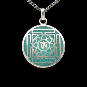 Op de afbeelding: Een zilveren ketting met een hanger in de vorm van een turquoise blauwe mandala met zilveren details. De hanger heeft een geometrisch patroon met een centraal symbool.