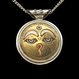 May include: Silver pendant necklace with a round, white and gold enamel design. The design features a stylized face with two eyes and a third eye in the center of the forehead.