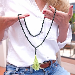 Puede incluir: Un collar de cuentas de color verde oscuro con un borla verde. El collar lo lleva una persona con una camisa blanca y pantalones vaqueros azules.