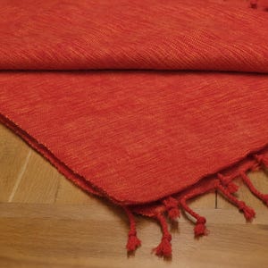 May include: A folded, textured, red woven textile with a fringe of knotted tassels. The fabric has a subtle, warm orange undertone and is displayed on a light brown wooden surface. The tassels are the same red color as the textile.